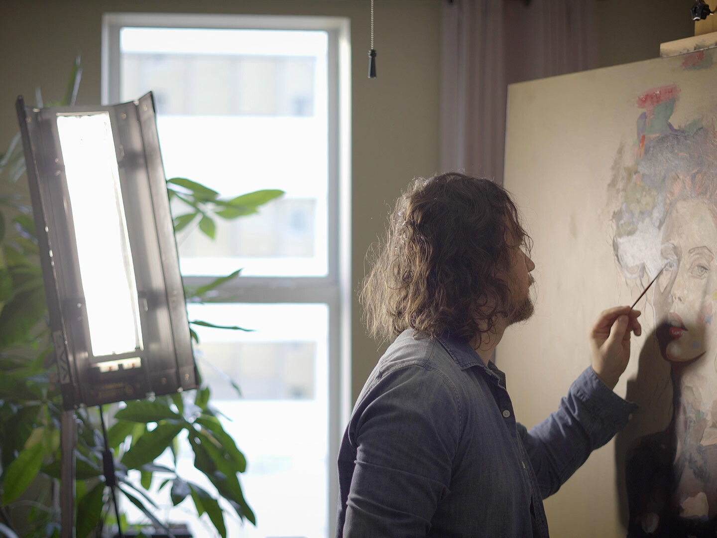 Artist at work on a painting of a woman's face, reminiscent of the Mona Lisa, in a sunlit studio.